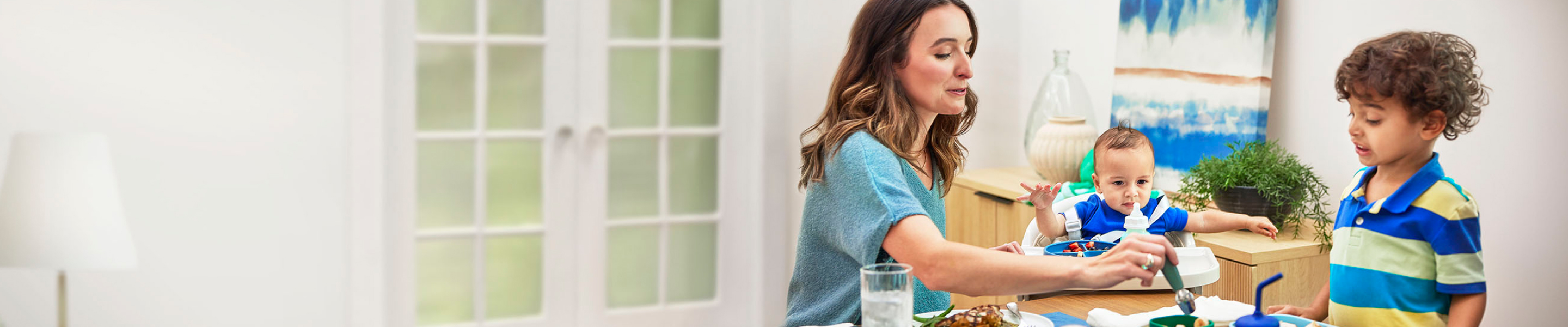 At dining table, mom is holding spoon in food for young son in striped shirt. A baby boy is between them in a high chair.