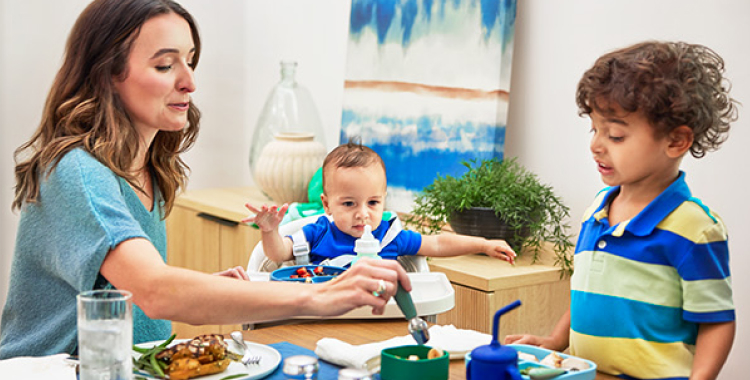 At dining table, mom is holding spoon in food for young son in striped shirt. A baby boy is between them in a high chair.