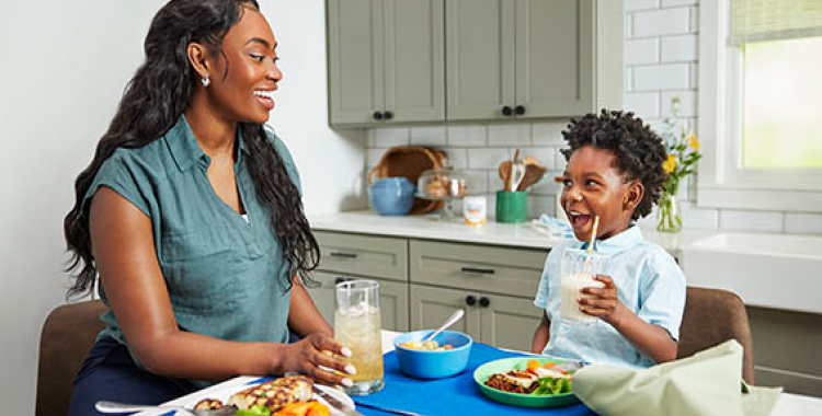 Young boy eating at the table in a bright kitchen, smiling and holding a glass of EleCare Jr. Mom is smiling back at him.