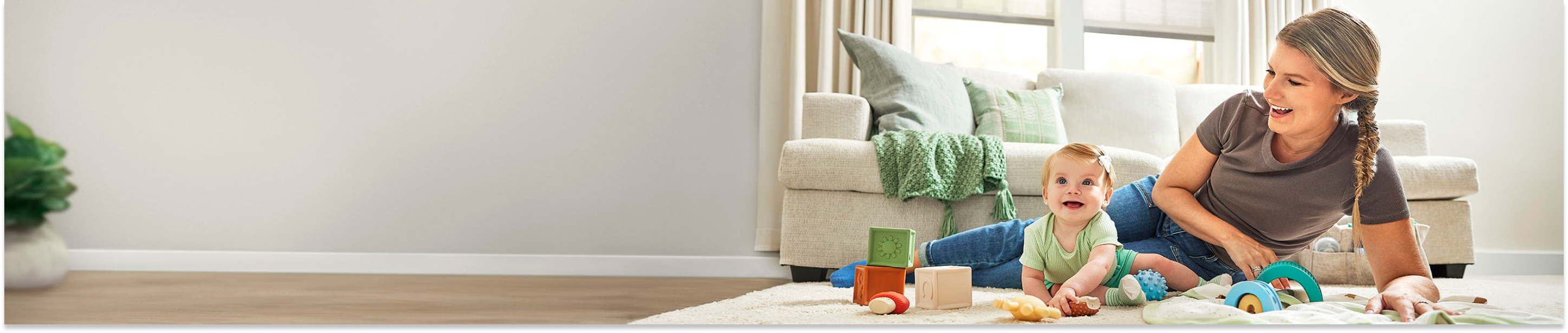 Mom in gray shirt and infant girl in green outfit playing with toys on a rug on the living room floor. Both are smiling.