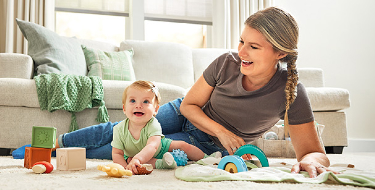 Mom in gray shirt and infant girl in green outfit playing with toys on a rug on the living room floor. Both are smiling.