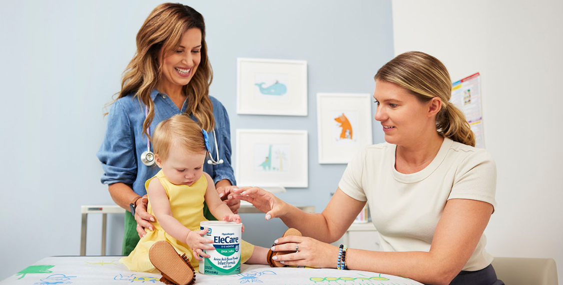 Doctor standing behind a baby girl, who is holding a can of EleCare Infant Formula. The baby’s mom is sitting next to them.