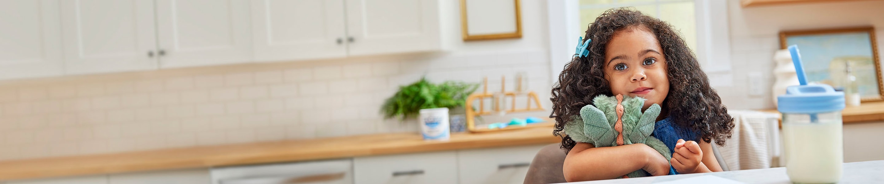 Young girl sitting at the kitchen table, holding a stuffed animal. A cup of EleCare Jr is in front of her. 