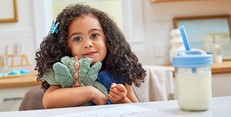 Young girl sitting at the kitchen table, holding a stuffed animal. A cup of EleCare Jr is in front of her. 