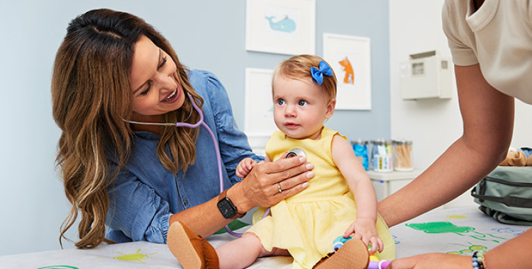 In a doctor’s office, a doctor is listening to a baby girl’s heart with a stethoscope. The girl is wearing a yellow dress.