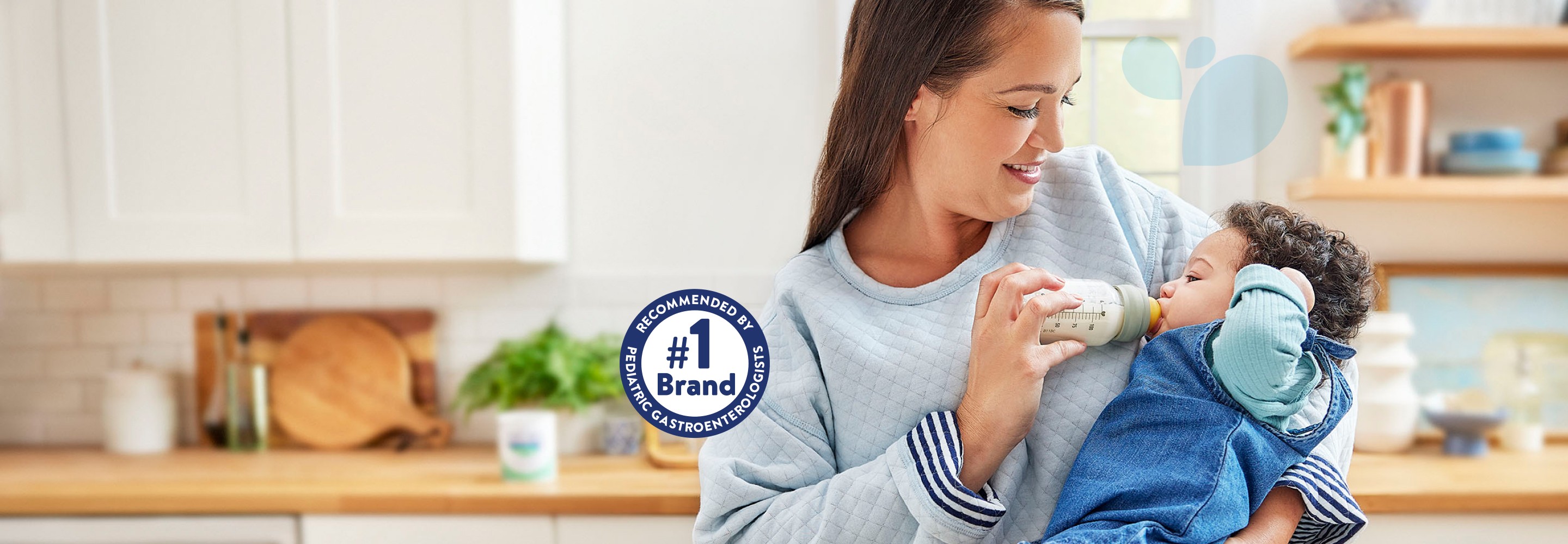 Mom standing in kitchen, holding her baby daughter, who is dressed in blue and being fed a bottle of EleCare.