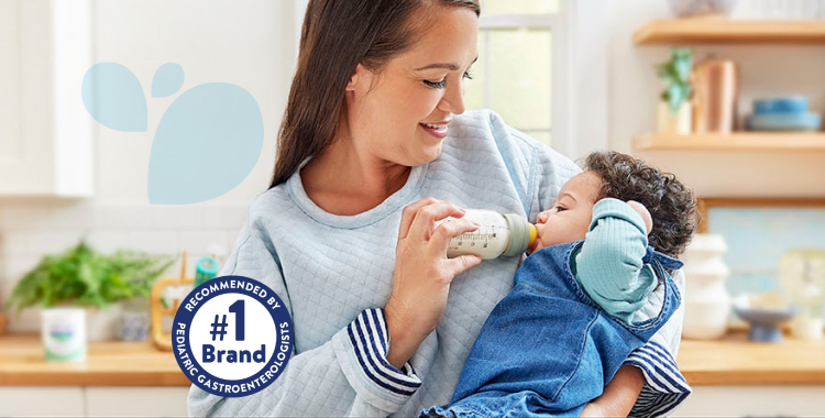 Mom standing in kitchen, holding her baby daughter, who is dressed in blue and being fed a bottle of EleCare.