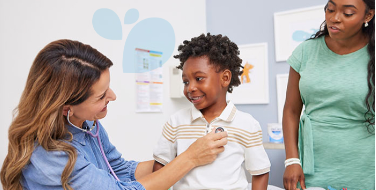 In a doctor’s office, a doctor is listening to a young boy’s heart with a stethoscope. His mom is standing next to him. 