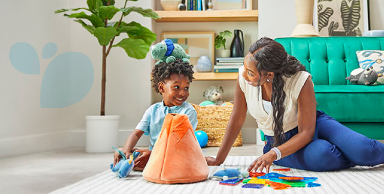 Woman sitting on the living room floor playing games with her young son, who has a stuffed animal on his head and in his hand.