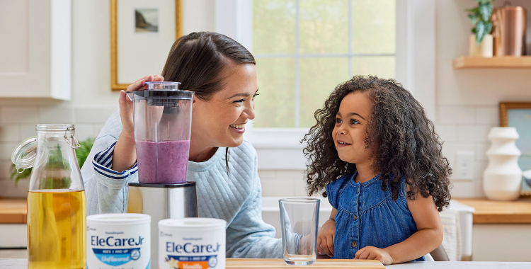 Woman in kitchen making a purple smoothie in a blender using EleCare Jr. Young girl watching and waiting for the smoothie.