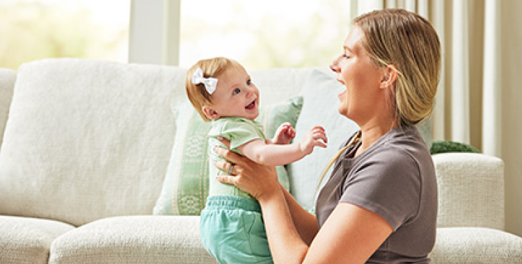 Woman in front of couch in living room, holding baby girl up; both are smiling at each other.