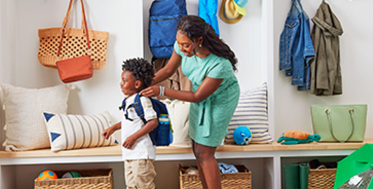 Woman in bright mudroom of house, placing blue backpack on young boy; both are smiling.