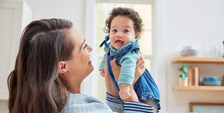 Woman standing in kitchen and smiling while holding up happy, smiling baby wearing blue