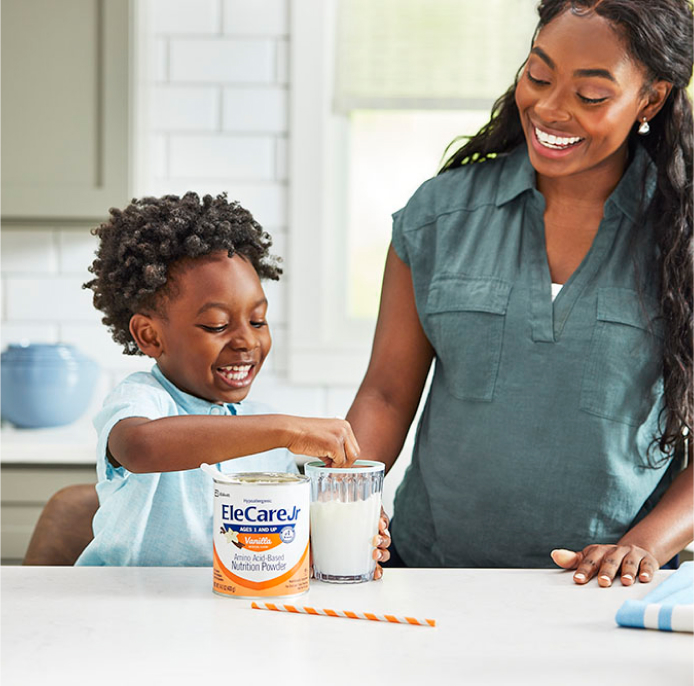 Young smiling boy sitting at the kitchen island and stirring a glass of EleCare Jr Vanilla, while his mom stands next to him.