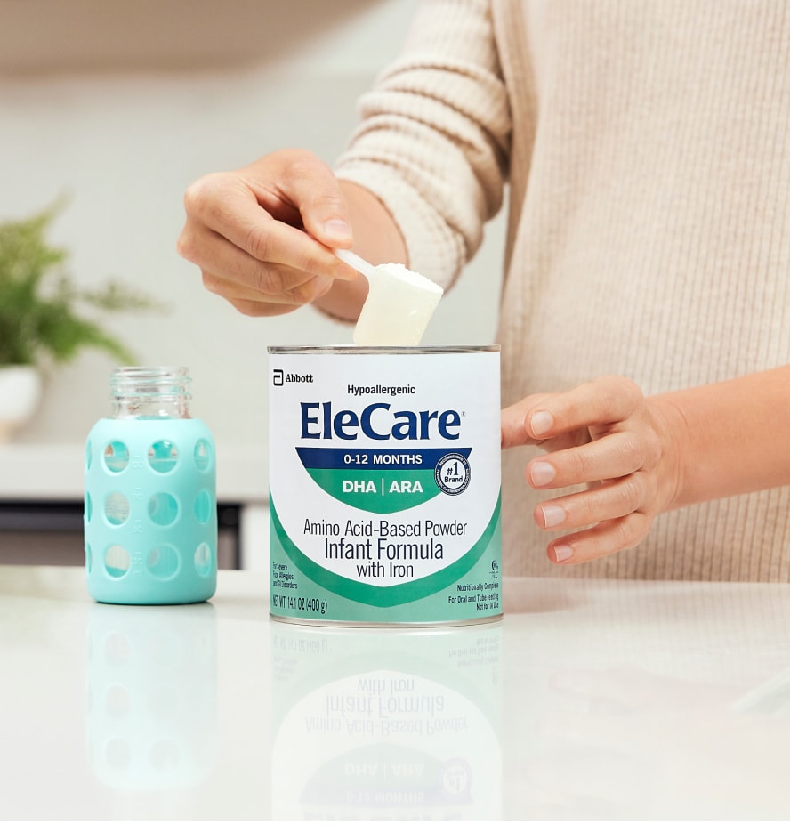 Close view of a woman in beige, in the kitchen, scooping EleCare Infant Formula from the can into a baby bottle with water.