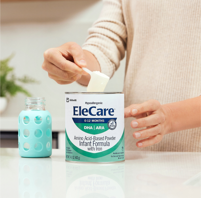 Close view of a woman in beige, in the kitchen, scooping EleCare Infant Formula from the can into a baby bottle with water.