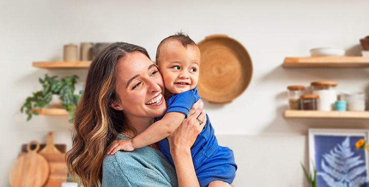 Woman holding her baby son, cheek to cheek, in the kitchen. Both are dressed in tones of blue and smiling.