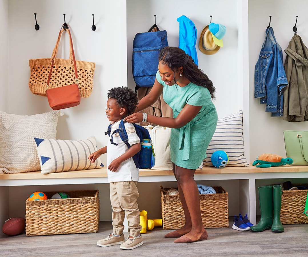 Woman in bright mudroom of house, placing blue backpack on young boy; both are smiling.