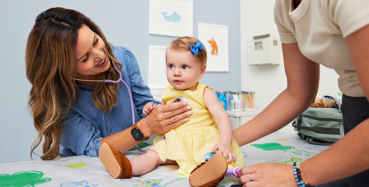 In a doctor’s office, a doctor is listening to a baby girl’s heart with a stethoscope. The girl is wearing a yellow dress.